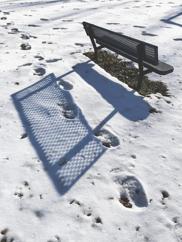 photo of an empty park bench casting a shadow on snowy ground