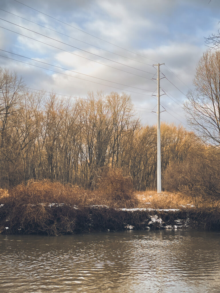 boring photo of a big boring steel utility pole along a boring brown river on a boring late winter day